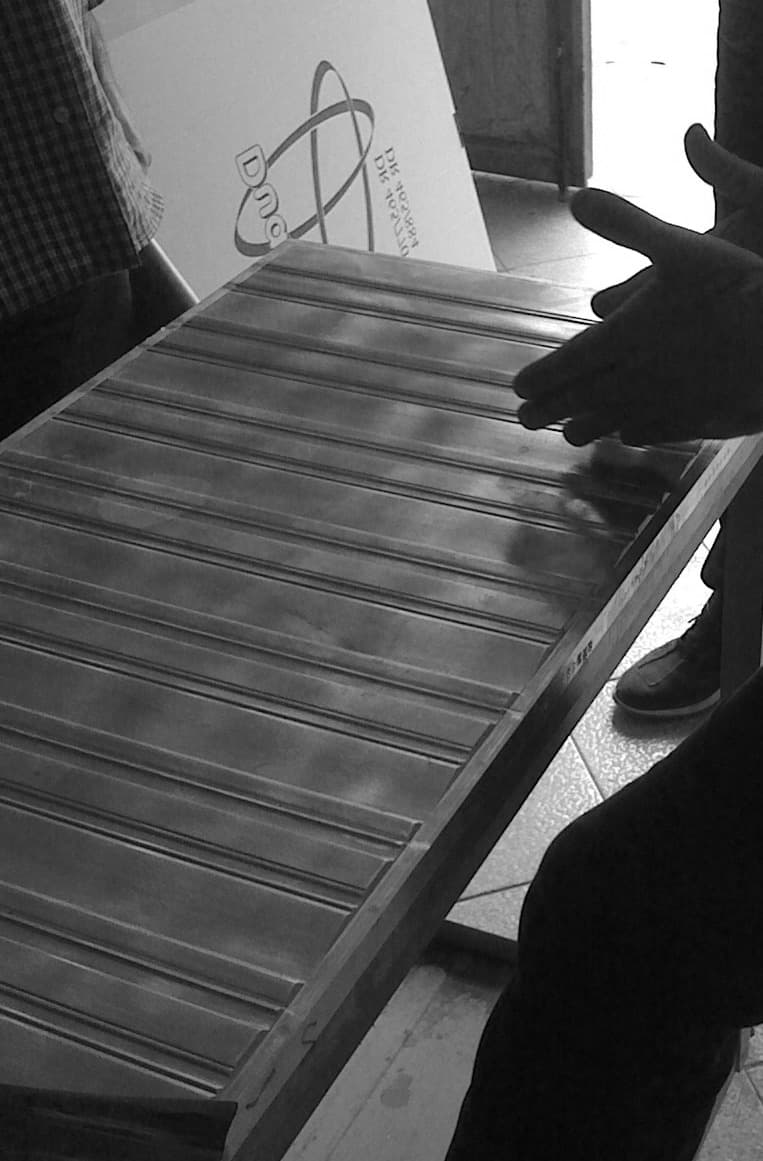 Close-up black and white view of a person with their hand suspended above a metal surface with parallel grooves. A sign with the "Dnc" logo can also be seen in the background.