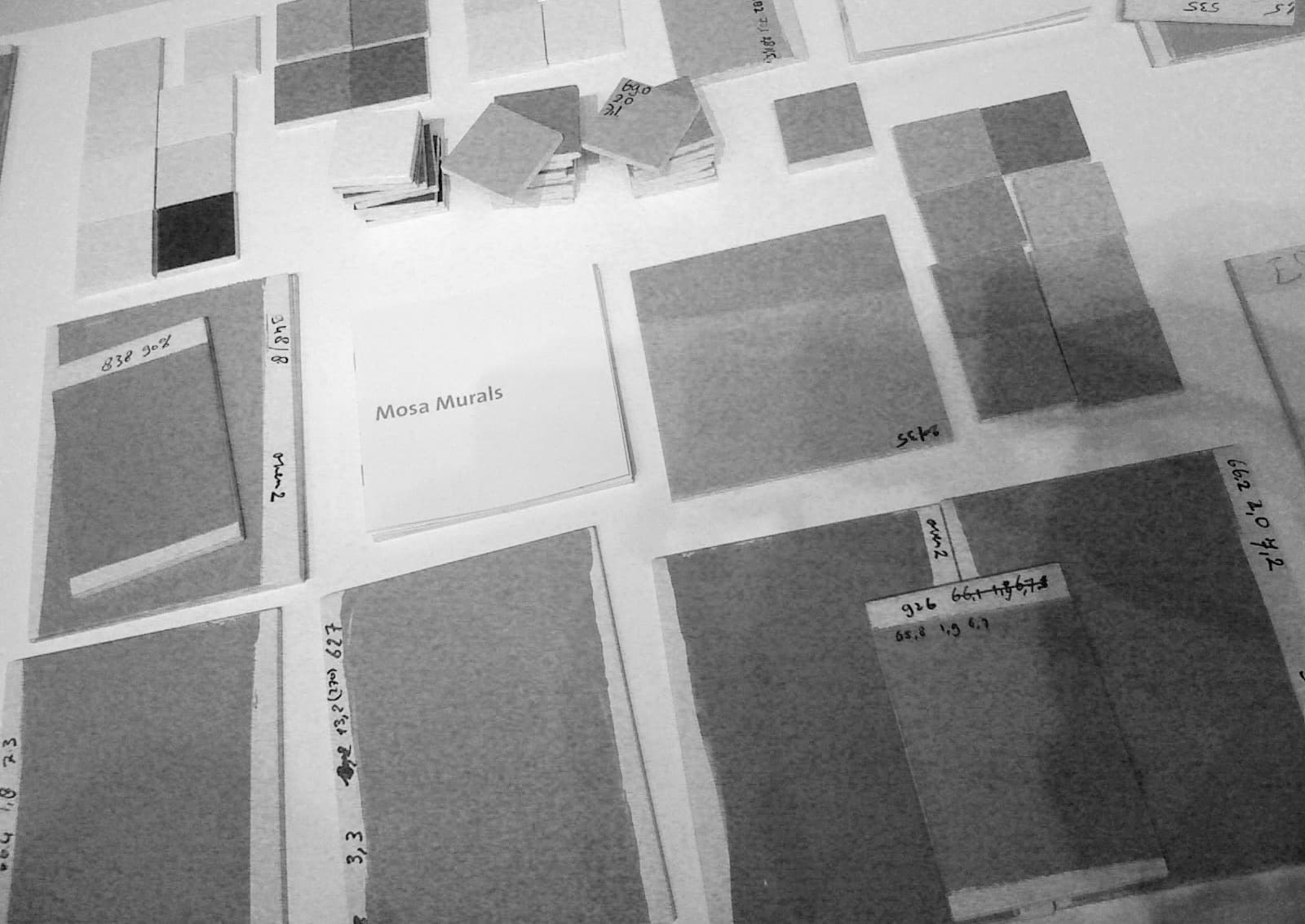 A view from above of a table or wall covered with numerous tile samples, data sheets, and color and handwritten notes, including a brochure titled "Mosa Murals." The image is black and white and features various shades of gray.