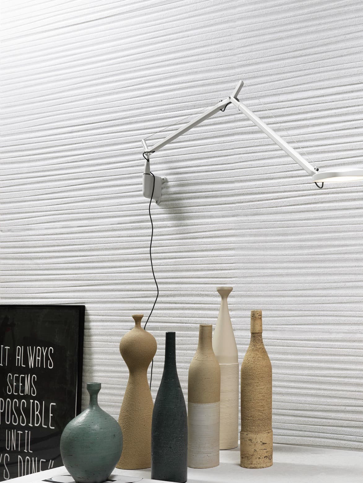 A close-up view of an interior. The back wall is covered with white relief tiles with a wavy pattern that creates a horizontal striped effect. A white wall lamp with an articulated arm,and is installed on the wall. Below, on a white shelf, several ceramic vases of various shapes and heights are arranged in a row, in shades of beige, brown, and blue-green. On the left side, a black frame with white text reads: "IT ALWAYS SEEMS IMPOSSIBLE UNTIL IT'S DONE."