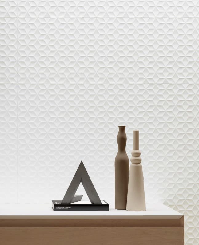 Una vista di un tavolo moderno in legno chiaro con una parete bianca sullo sfondo. Il muro è rivestito con piastrelle bianche tridimensionali che presentano un motivo geometrico a triangoli e cubi in rilievo. Sul tavolo, due vasi di ceramica di forma allungata, uno marrone e uno bianco, sono posizionati accanto a una scultura astratta in cemento a forma di "A".