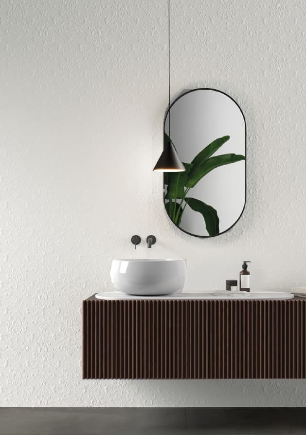 Un lavabo moderno con una ciotola bianca a forma di semisfera appoggiata su un mobile sospeso con una superficie a stecche di legno scuro. Sul muro a destra, un grande specchio ovale con una cornice nera riflette una pianta d'appartamento. Il muro di fondo, in piastrelle bianche, ha una texture a rilievo e un effetto ruvido. Una lampada a sospensione nera pende sopra il lavabo.