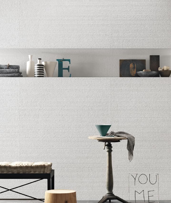 Un'immagine di una parete bianca con una texture a rilievo che ricorda un tessuto a trama fitta. La parte superiore presenta una nicchia orizzontale incassata nel muro, utilizzata come mensola per oggetti decorativi come vasi, libri, ciotole e la scultura "E" in metallo azzurro. La parte inferiore dell'immagine mostra una panca in tessuto peloso, un piccolo sgabello cilindrico in legno e un tavolino nero con una ciotola verde. Un'installazione artistica che forma le parole "YOU ME" è in primo piano.
