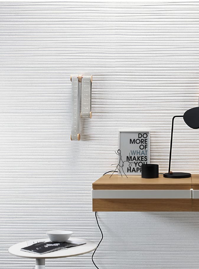 Una parete bianca con una texture orizzontale a rilievo che richiama l'effetto di un tessuto ondulato. Al centro della parete è appeso un calendario in legno e tessuto. In primo piano, una scrivania moderna in legno chiaro e bianco è parzialmente visibile, con una lampada da tavolo nera e un piccolo quadro con la scritta "DO MORE OF WHAT MAKES YOU HAPPY".