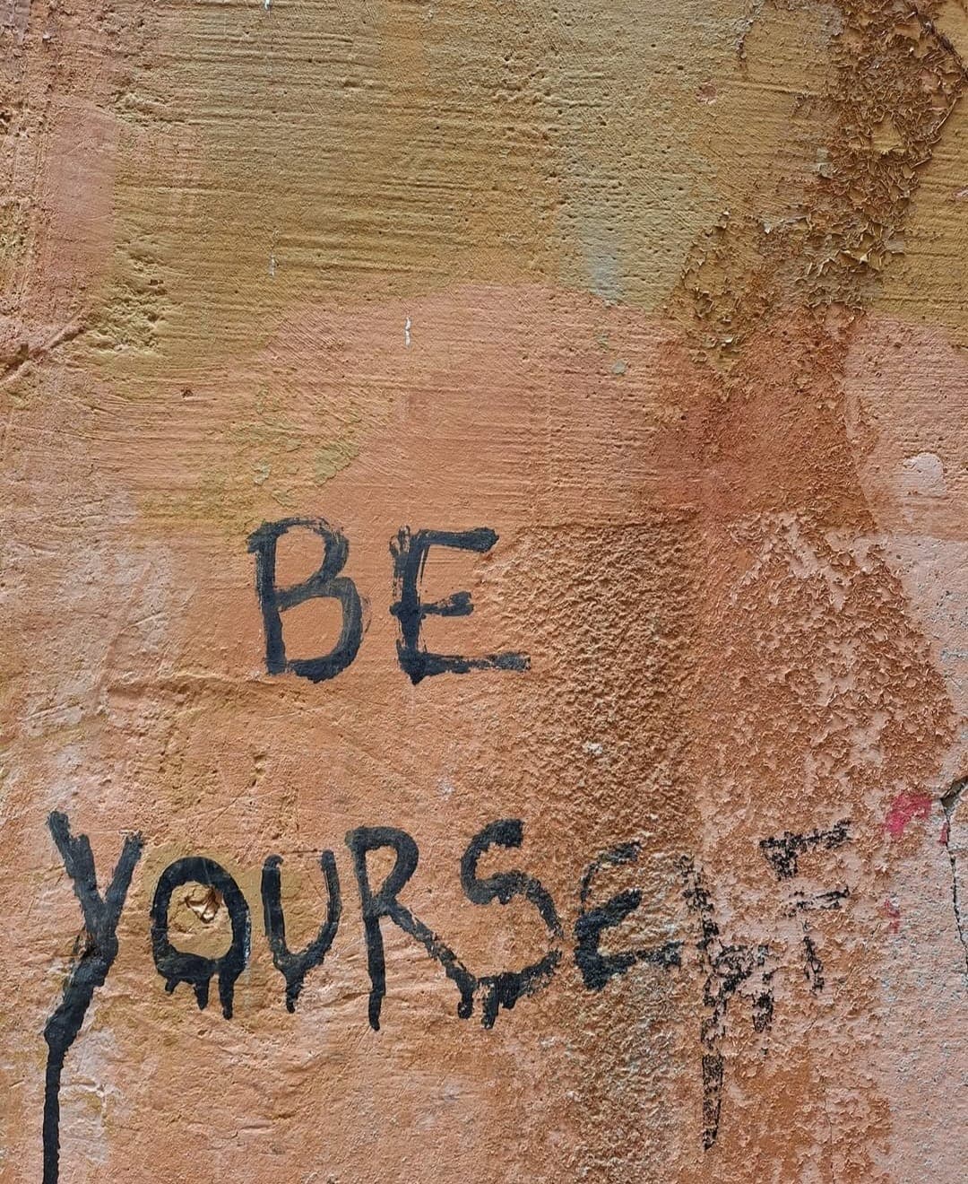 A close-up of a worn, peeling wall, with colors ranging from orange to brown to beige. In the foreground, the words "BE YOURSELF" are hand-painted in black, with a few drops of paint dripping from the letter "E" in "YOURSELF".
