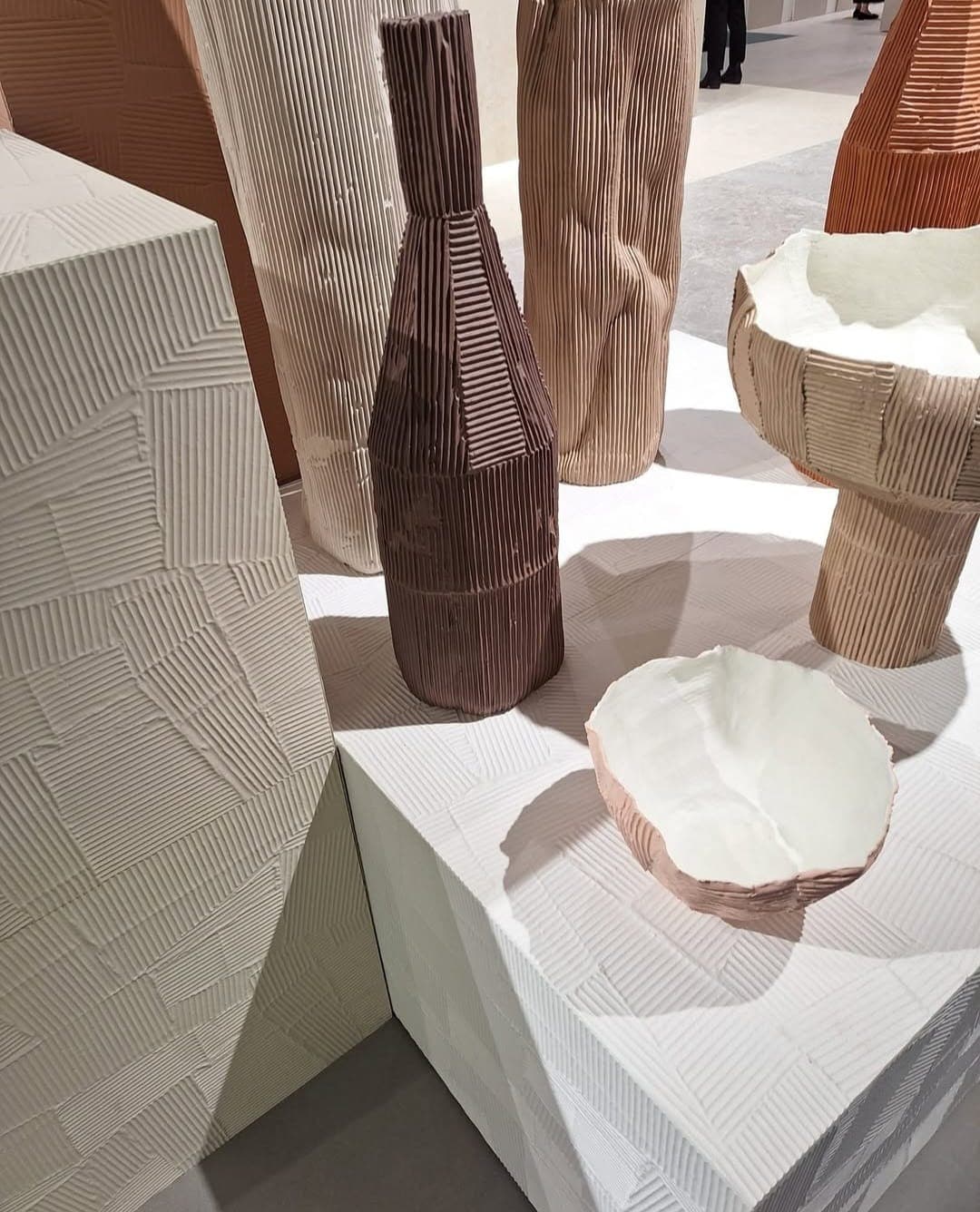An arrangement of ceramic vases and sculptures in a minimalist room. The works, ranging from beige to dark brown, feature a relief pattern resembling vertical striations or a grid-like texture. In the foreground, a white sculpture in the shape of a broken bowl is positioned on a white display block with a grid-like relief texture.